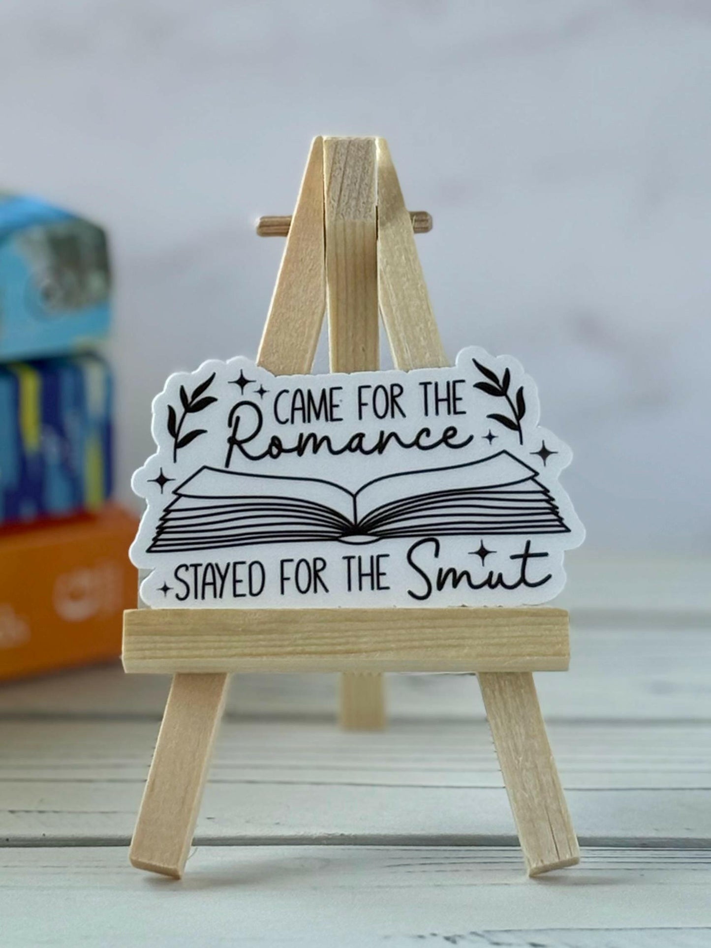Black text on a white sticker that says “Came for the romance, stayed for the smut,” displayed on a bookish surface