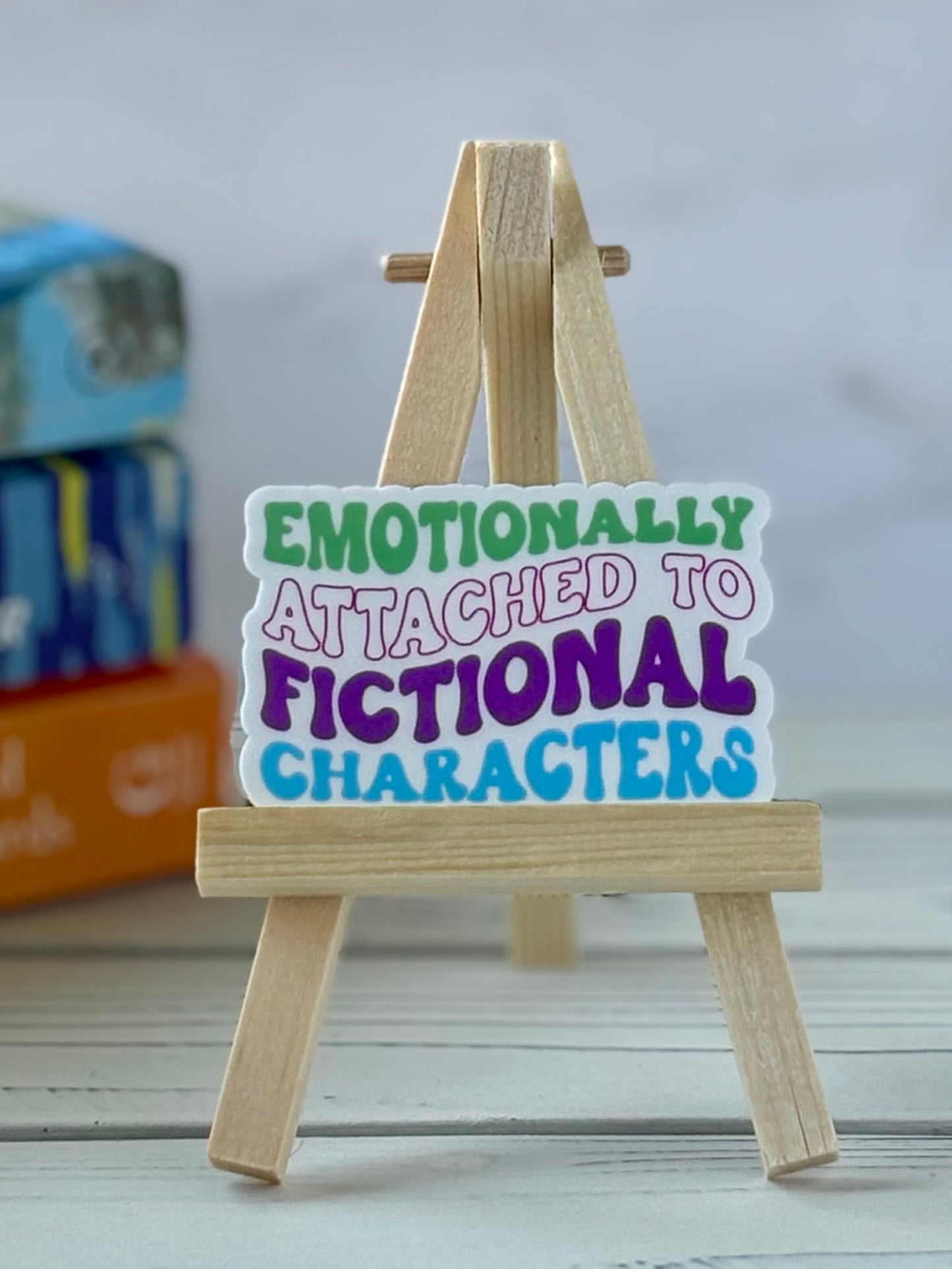 Wooden easel with a vinyl sticker reading 'Emotionally Attached to Fictional Characters' on a blurred background.