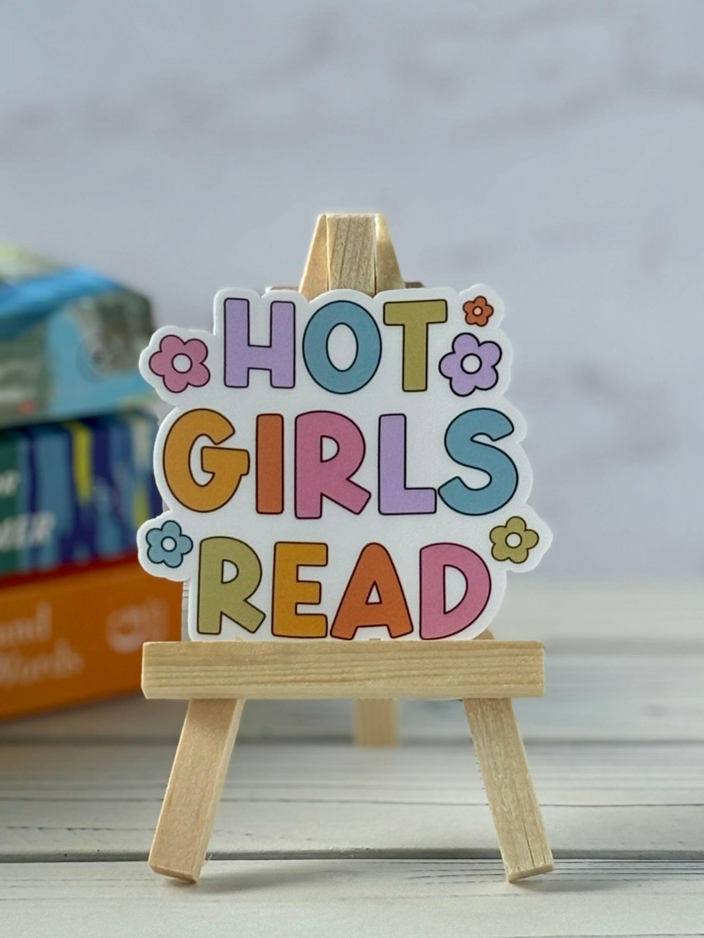 Retro-style vinyl sticker that says “Hot Girls Read” in multi-color bubble letters with flower accents, shown on a small wooden easel.