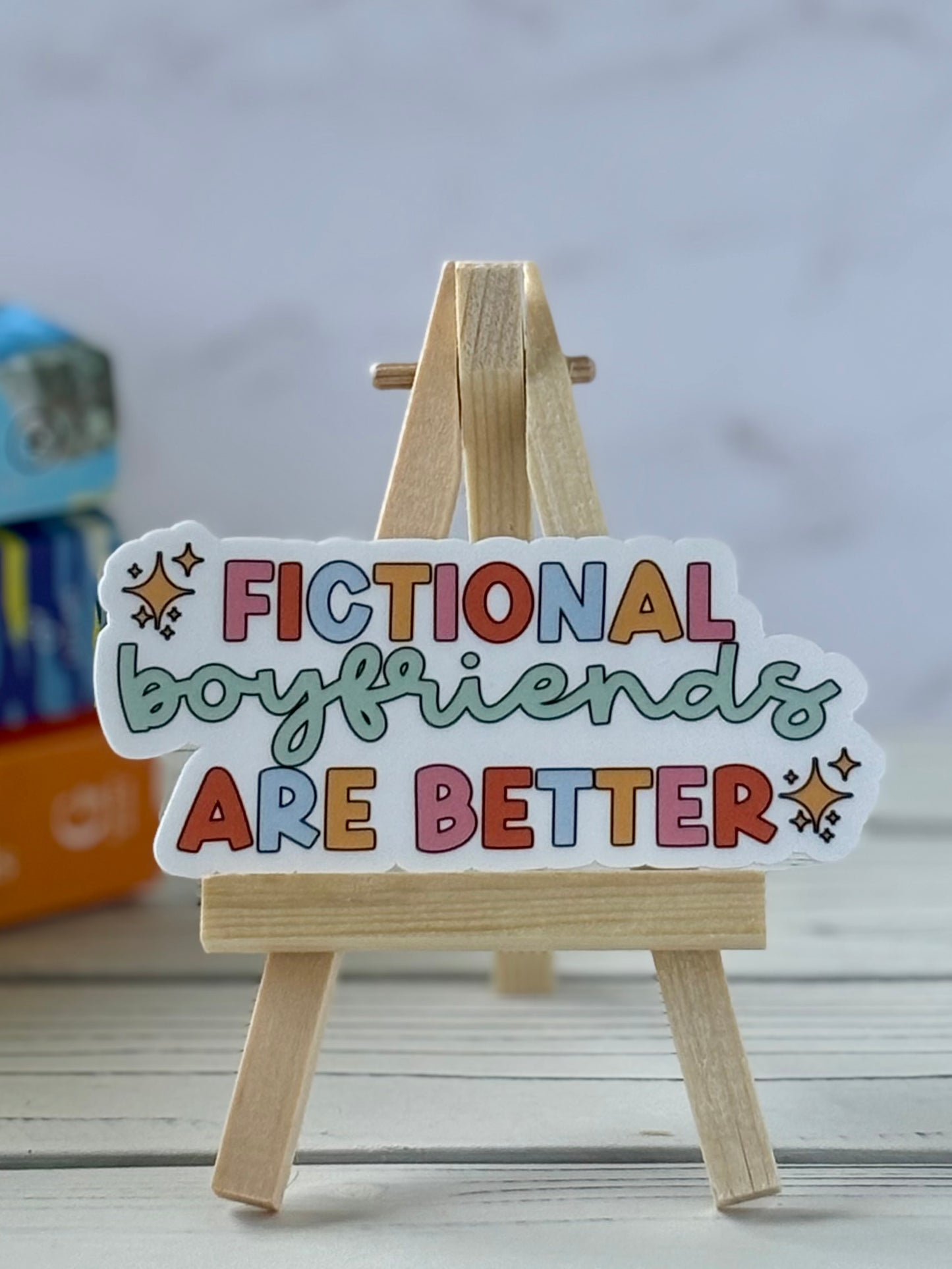 Sticker with 'Fictional boyfriends are better' text on a wooden stand against a blurred background
