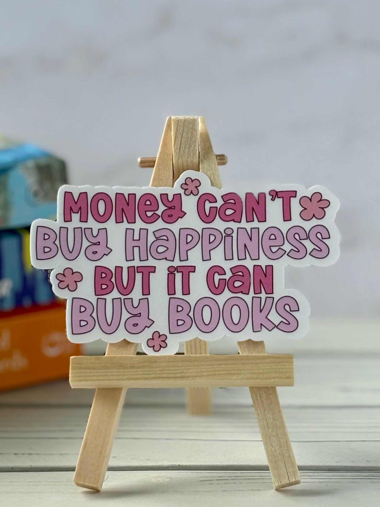 Retro bookish sticker that says “Money Can’t Buy Happiness But It Can Buy Boks” in pink text with flower accents, displayed on a mini easel.