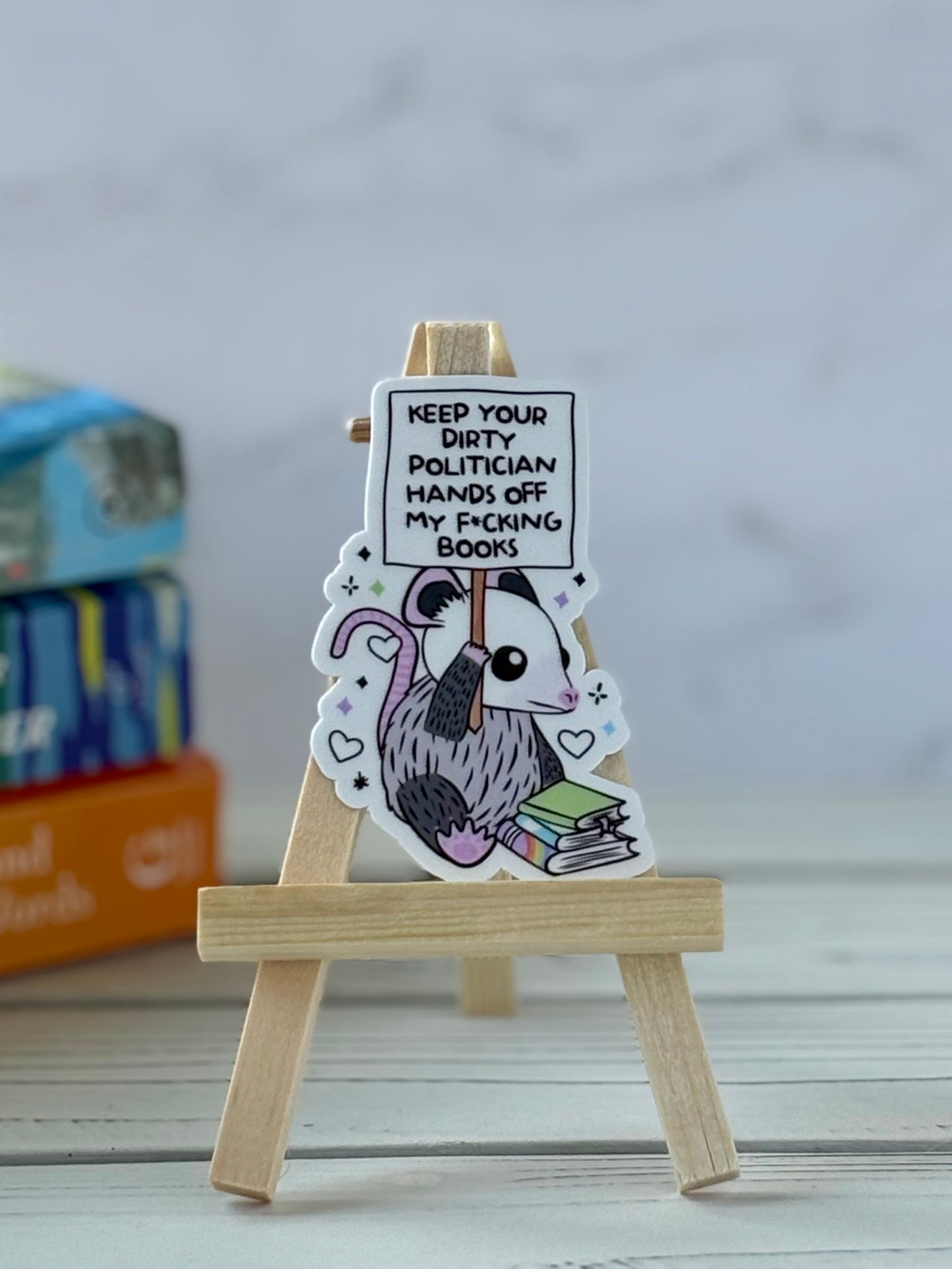 Sticker of an opossum holding a protest sign reading “Keep Your Dirty Politician Hands Off My F*cking Books” behind a stack of books
