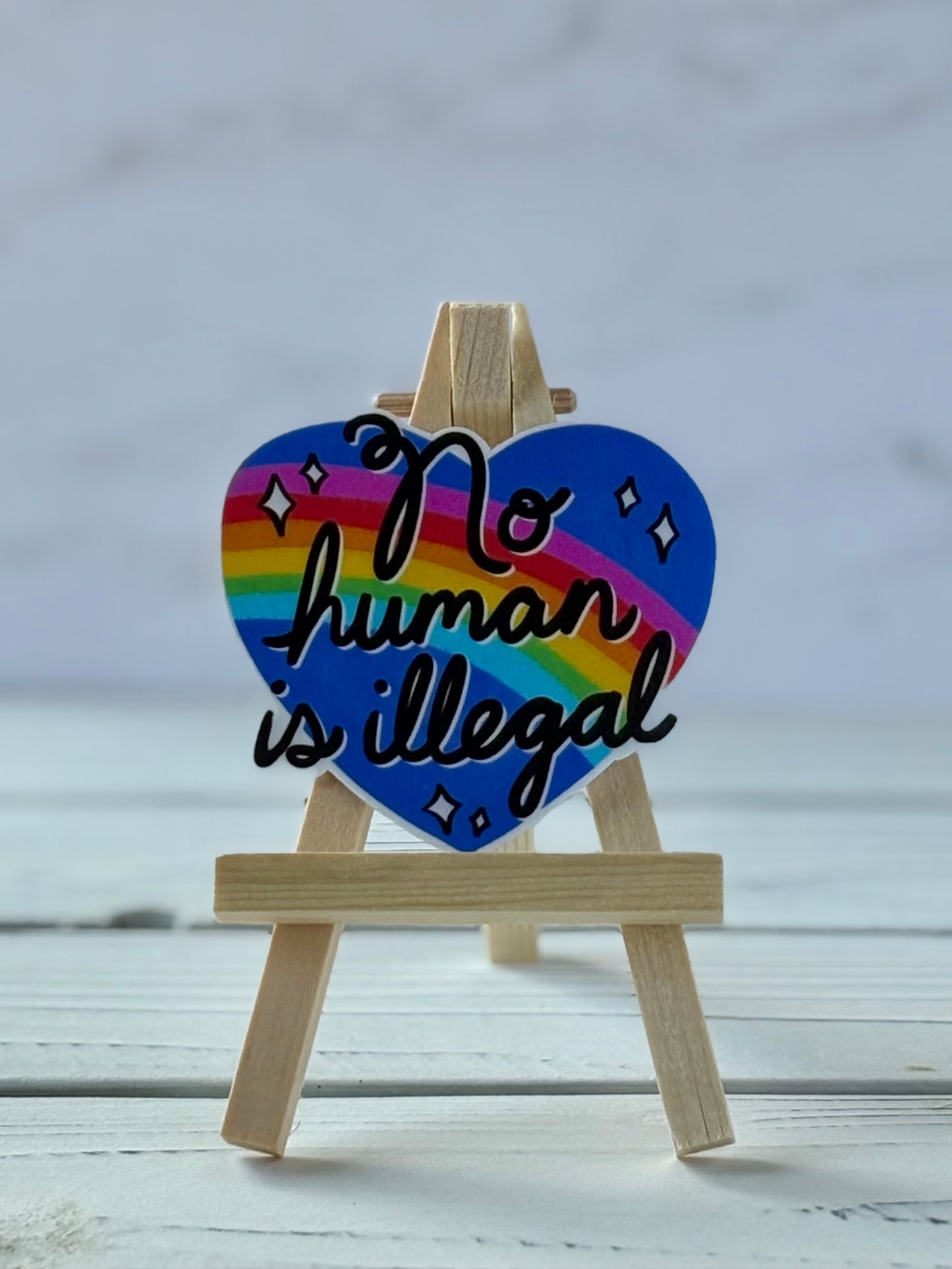 Sticker with black script text “No Human Is Illegal” on a blue heart with a rainbow