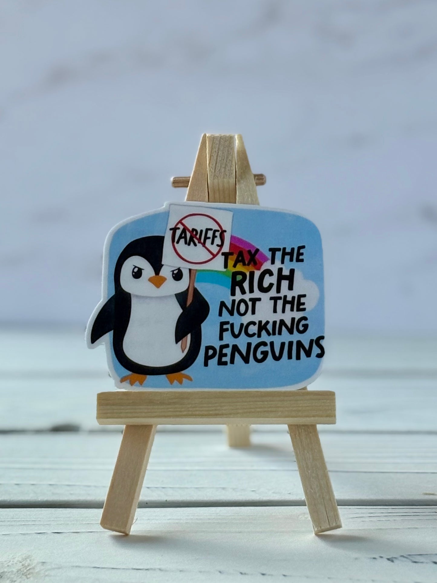 Sticker with penguin holding a crossed-out “tariffs” sign; rainbow background with black text “Tax the rich not the f*cking penguins”