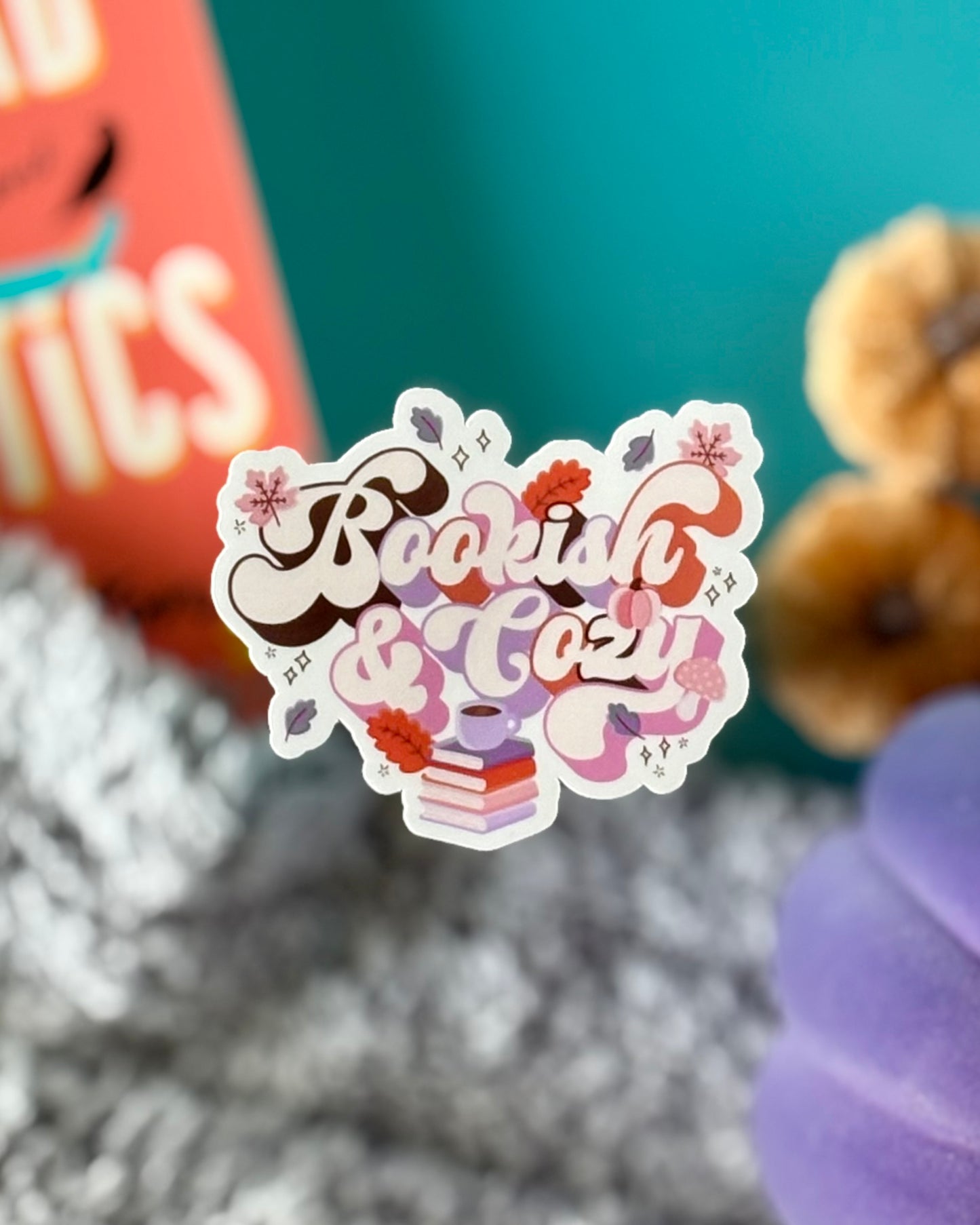 Set of a retro Bookish & Cozy bookmark and matching vinyl sticker, both with brown, purple, orange, and pink text, coffee-topped books, pumpkins, leaves, and mushrooms.
