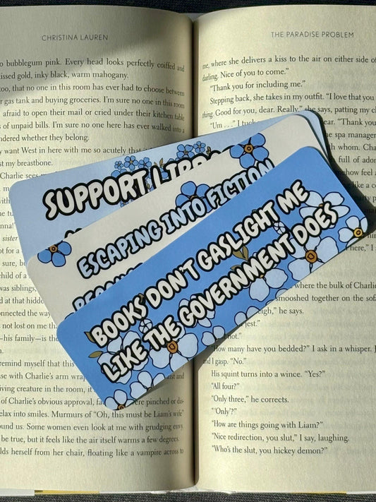 Blue floral bookmarks with political messages: “Books Don’t Gaslight Me Like The Government Does,” “Support Libraries, Not Billionaires,” and “Escaping Into Fiction Because Reality Is Too Red”