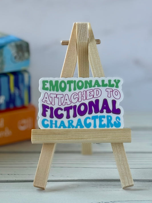 Wooden easel with a vinyl sticker reading 'Emotionally Attached to Fictional Characters' on a blurred background.