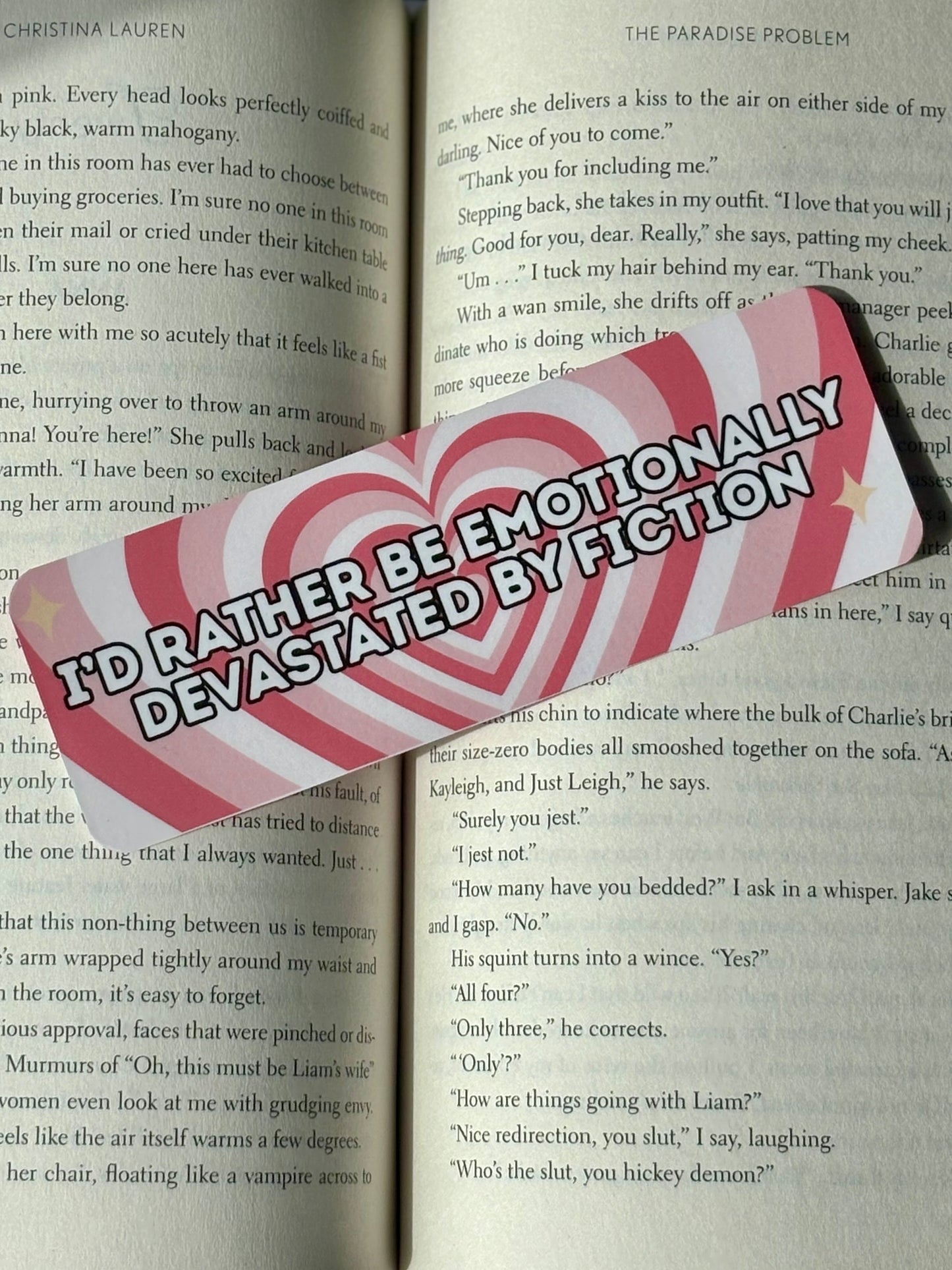 Bookmark with concentric hearts behind text that says “I’d Rather Be Emotionally Devastated By Fiction” on an open book.