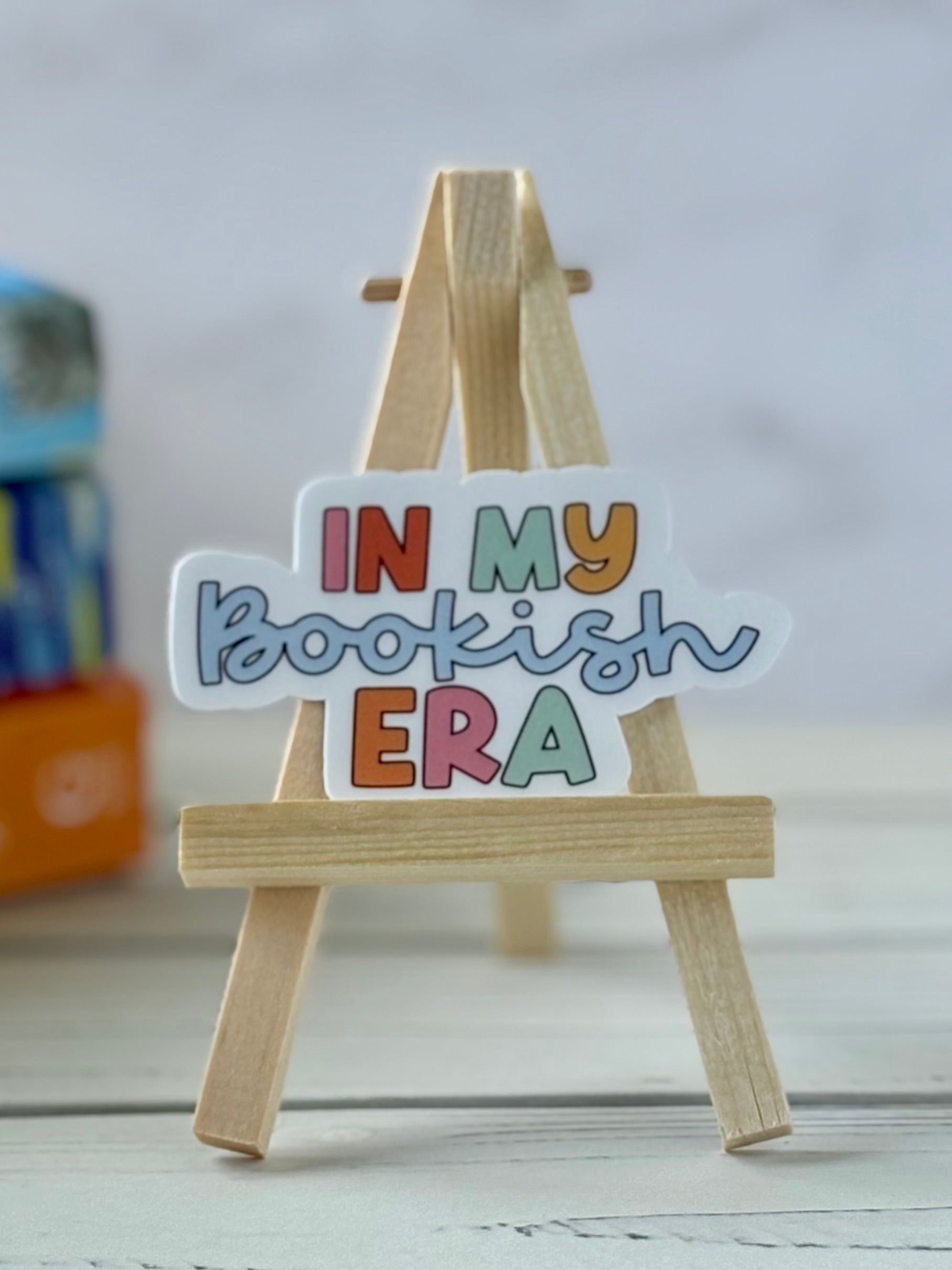 Small wooden easel with a colorful sign reading 'IN MY Bookish ERA' on a light background.