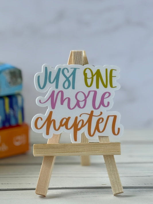 Colorful 'Just One More Chapter' sticker on a wooden stand with a blurred background