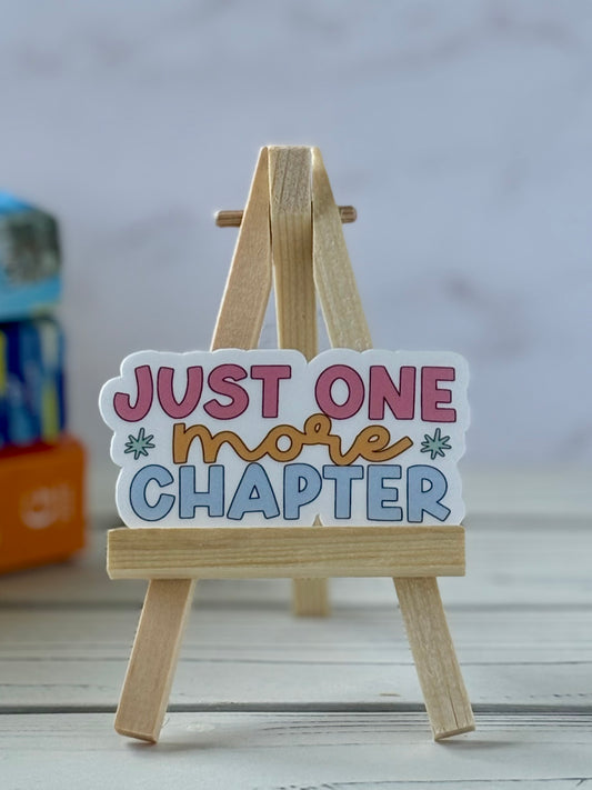 Small wooden easel with a colorful sign reading 'Just One More Chapter' on a neutral background.