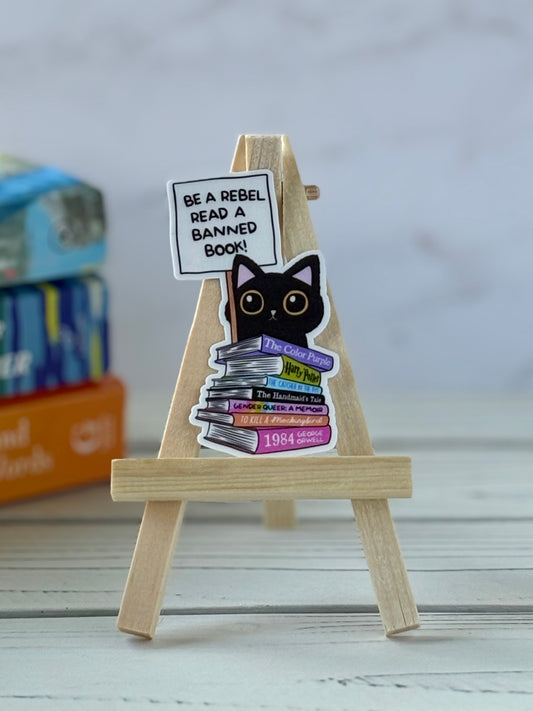 Sticker of a black cat holding a “Be a Rebel Read a Banned Book!” sign behind a stack of banned books