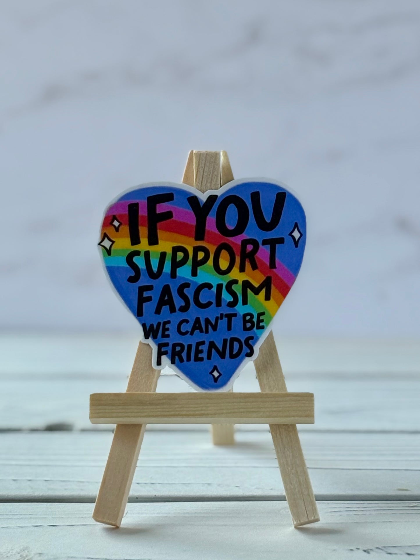 Sticker with black text “If You Support Fascism We Can’t Be Friends” on blue heart with rainbow overlay