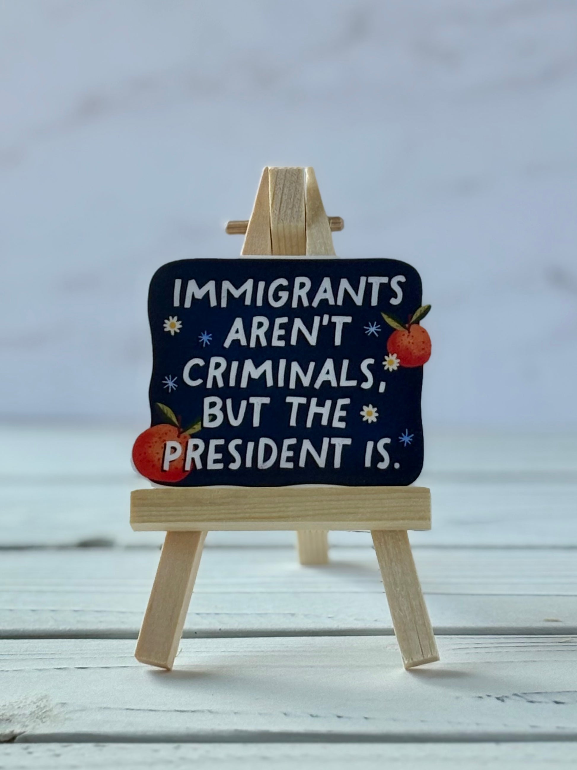 Sticker with white text reading “Immigrants Aren’t Criminals, But the President Is” on navy background with daisies, oranges, and dandelion seeds