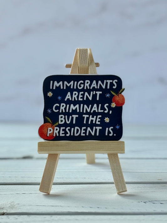 Sticker with white text reading “Immigrants Aren’t Criminals, But the President Is” on navy background with daisies, oranges, and dandelion seeds