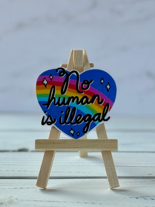 Sticker with black script text “No Human Is Illegal” on a blue heart with a rainbow