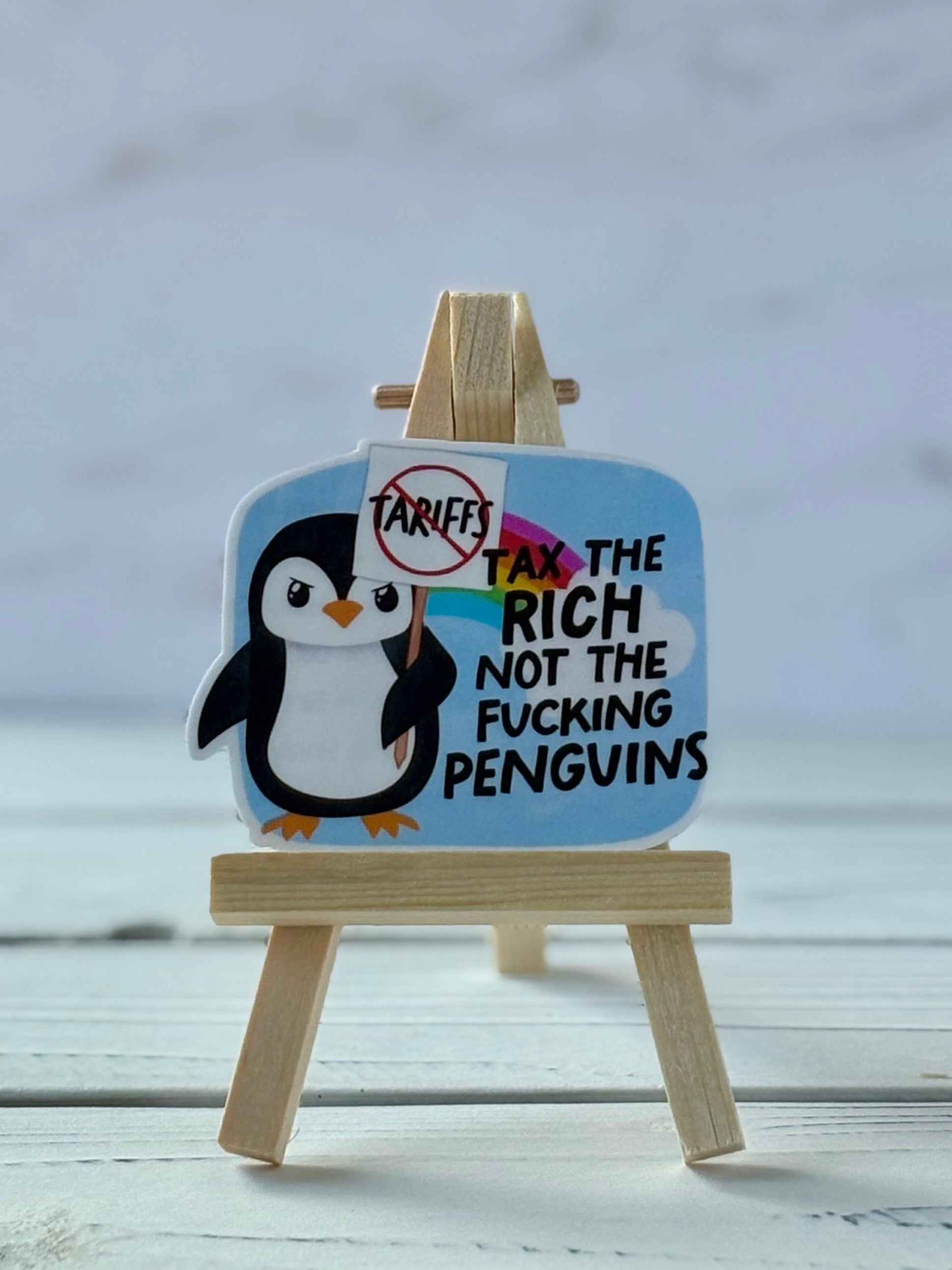 Sticker with penguin holding a crossed-out “tariffs” sign; rainbow background with black text “Tax the rich not the f*cking penguins”