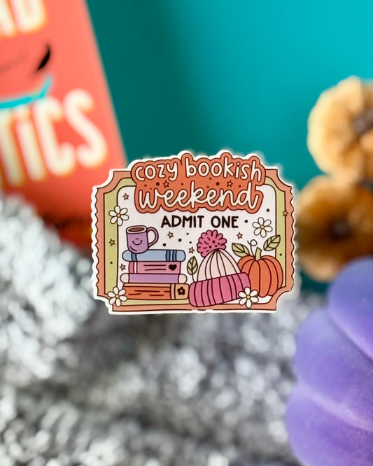 Retro ticket sticker reading “Cozy Bookish Weekend Admit One” with book stack, coffee mug, pumpkin, and pink beanie in fall colors