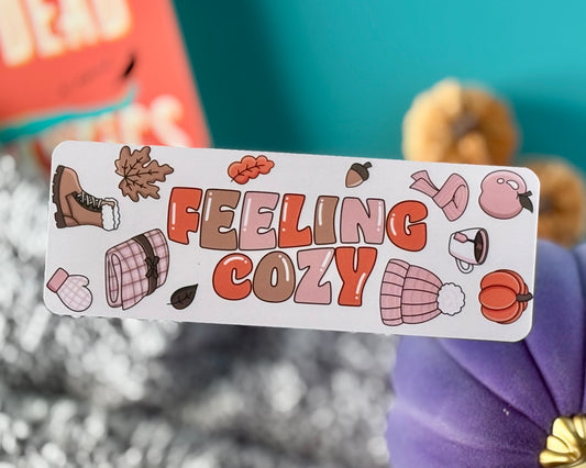 Bookmark with retro text “Feeling Cozy” in warm fall tones, surrounded by boots, beanies, pumpkins, mugs, leaves, and acorns