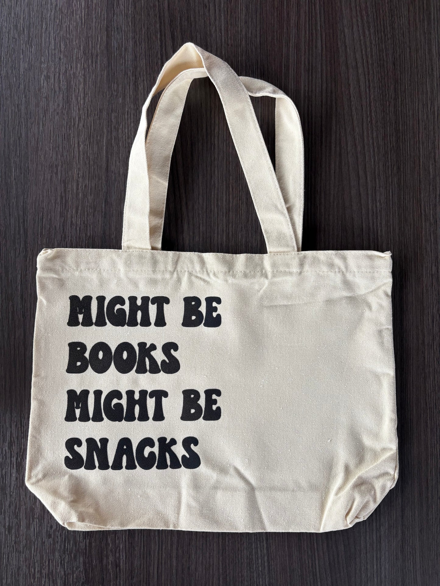 Flat lay of cute bookish tote that says Might Be Books Might Be Snacks