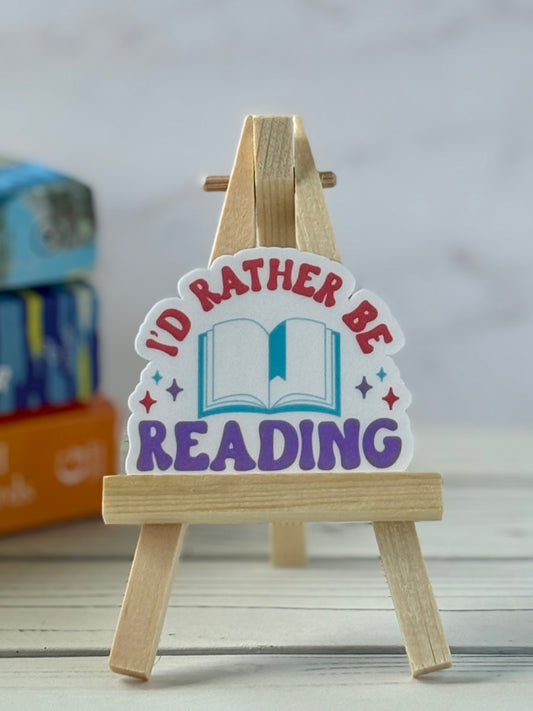 Sticker with 'I'd Rather Be Reading' text on a wooden stand against a neutral background